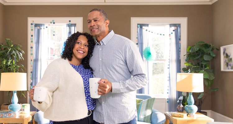 A man and a woman are standing, happily drinking coffee in their home.